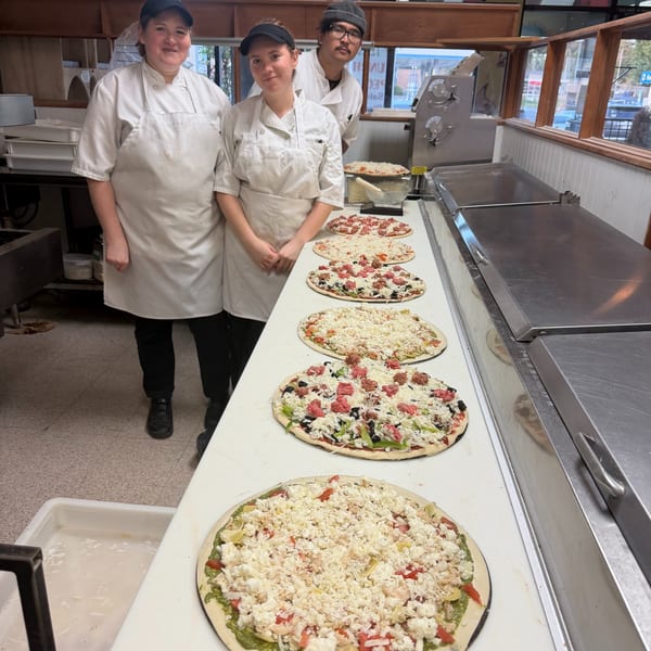 Three Cerello's Pizza chefs stand behind a counter with multiple uncooked pizzas topped with cheese and ingredients.